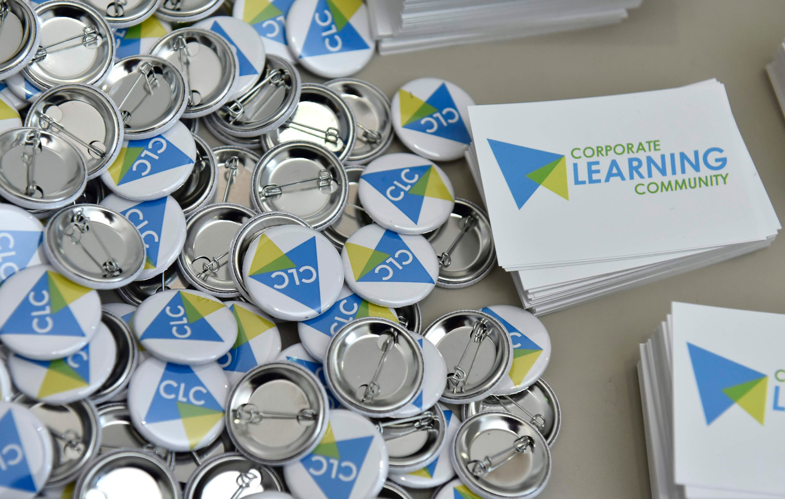 CLC Buttons und Sticker auf dem #CLC19HH, Foto von Christian Augustin, im Auftrag von Nordmetall, CC BY 4.0, Auf dem Bild sieht man viele runde Ansteck-Buttons. Auf den Buttons
steht „CLC" und es sind bunte Dreiecke, das Logo der CLC, zu sehen.
Neben den Buttons liegen kleine Karten. Auf den Karten steht: „Corporate
Learning Community". 
