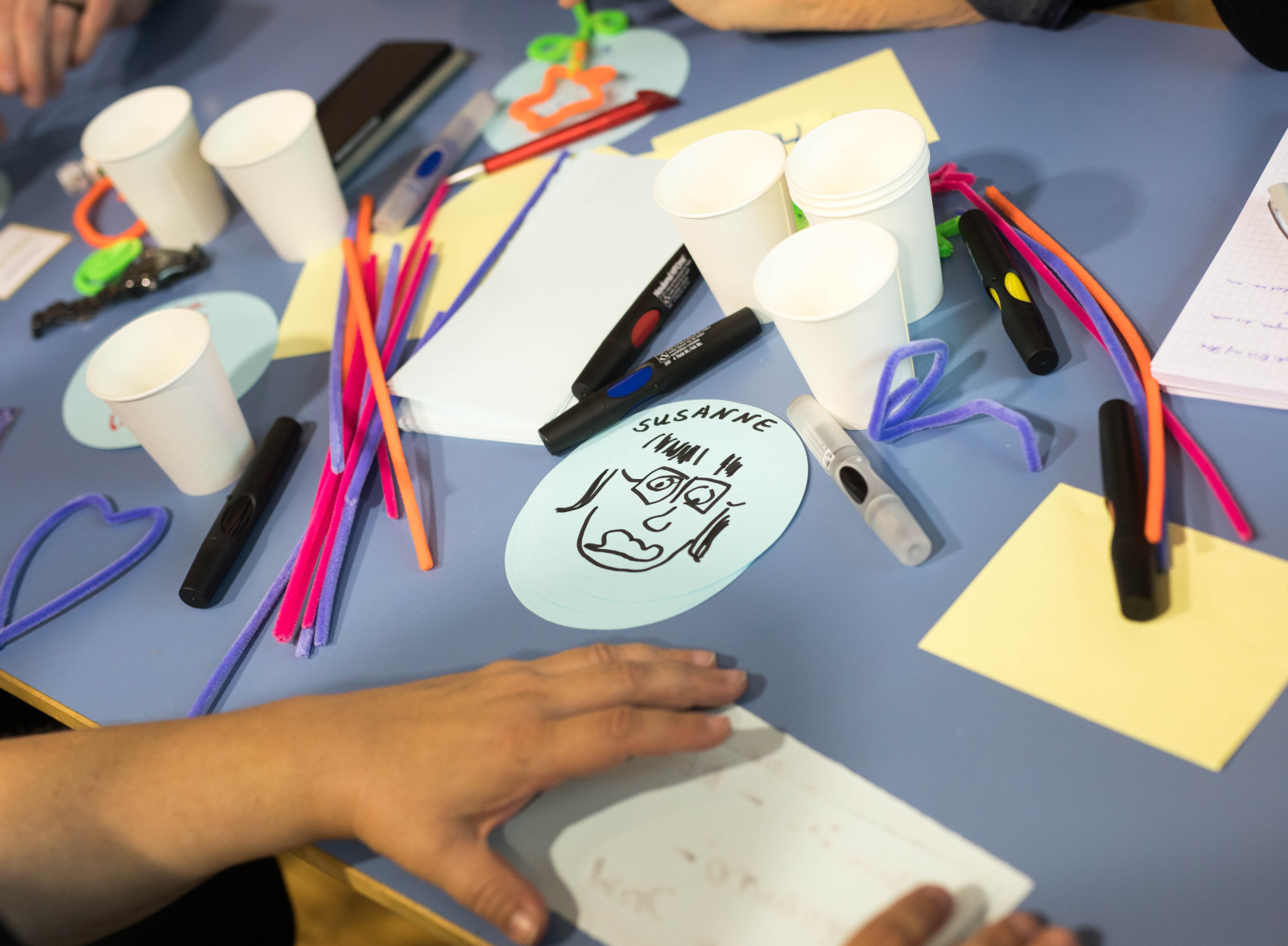 Barcamp-Sessiongestaltung auf dem #CLC17 (Foto von Frank Rumpenhorst), Das Bild stammt vom Corporate Learning Camp 2017. Auf dem Bild sieht man einen Basteltisch mit vielen bunten Materialien: Pfeifenreiniger, Stifte, Papier, Scheren und Becher. In der Mitte liegt ein ovaler Zettel mit einer gezeichneten Figur und dem Namen „SUSANNE". Mehrere Personen arbeiten gemeinsam am Tisch. Die Stimmung wirkt kreativ und spielerisch - es scheint eine Gruppenübung oder ein Einstieg in einen Workshop zu sein.