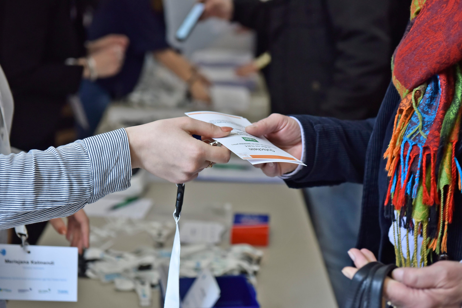 Check In auf dem CLC19HH (Foto von Christian Augustin, im Auftrag von Nordmetall, CC BY 4.0), Das Foto stammt vom Corporate Learning Camp 2019 in Hamburg. Das Bild zeigt die Anmeldung bei der Veranstaltung. Eine Person übergibt einem Gast einen Namensausweis mit Schlüsselband. Auf dem Tisch liegen weitere Namensschilder. Im Hintergrund sind weitere Personen zu sehen.