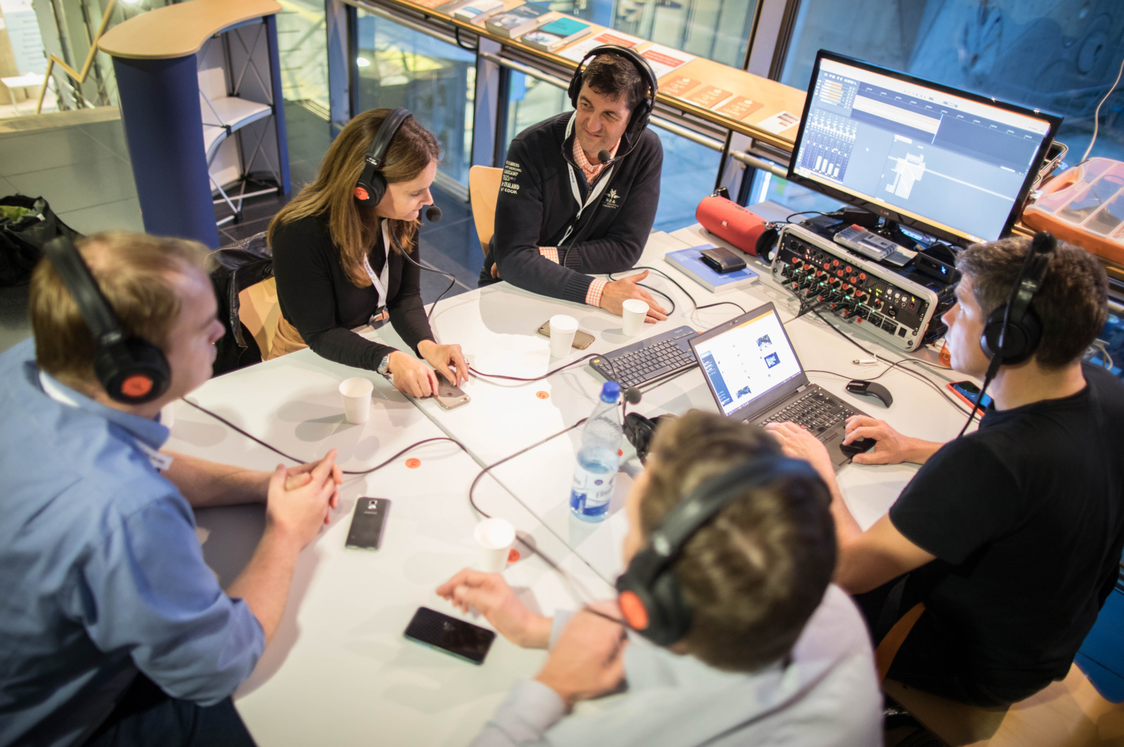 Podcast-Aufnahme auf dem CLC17 (Foto von Frank Rumpenhorst), Das Foto stammt vom Corporate Learning Camp 2017. Fünf Personen sitzen an einem Tisch und tragen große Kopfhörer mit Mikrofonen. Sie sprechen miteinander und schauen konzentriert. Auf dem Tisch stehen Laptops, eine Wasserflasche und Technik zur Tonaufnahme. Das Bild zeigt eine Podcast- oder Radioaufnahme. 