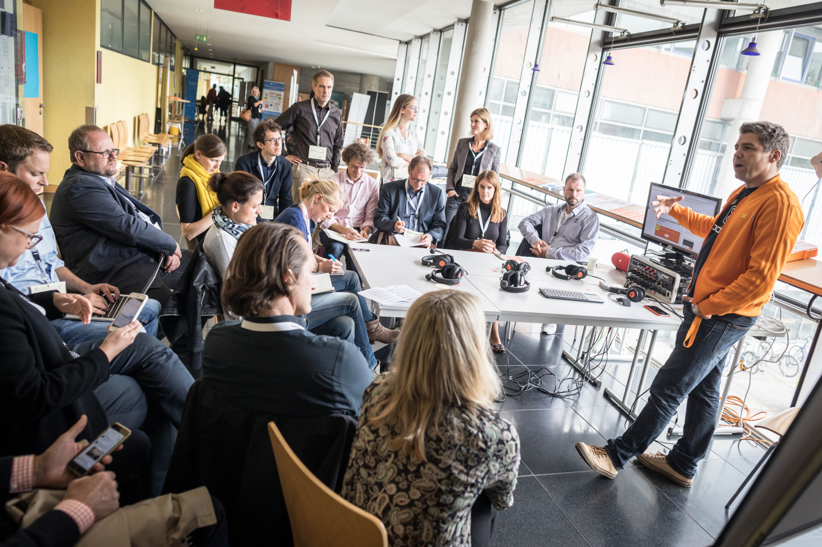 Session auf dem #CLC17 (Foto von Frank Rumpenhorst), Das Bild stammt vom Corporate Learning Camp 2017. Auf dem Bild sieht
man eine Gruppe von Menschen, die in einem offenen Raum zusammensitzen.
Sie hören einer Person, Simon Dückert, zu, die vorne steht und etwas
erklärt. Die Person trägt eine orange Jacke und zeigt auf einen
Bildschirm. Auf dem Tisch liegen Mikrofone und Kopfhörer. Einige
Teilnehmende machen sich Notizen oder schauen auf ihre Handys. Die
Stimmung wirkt aufmerksam und
interessiert.