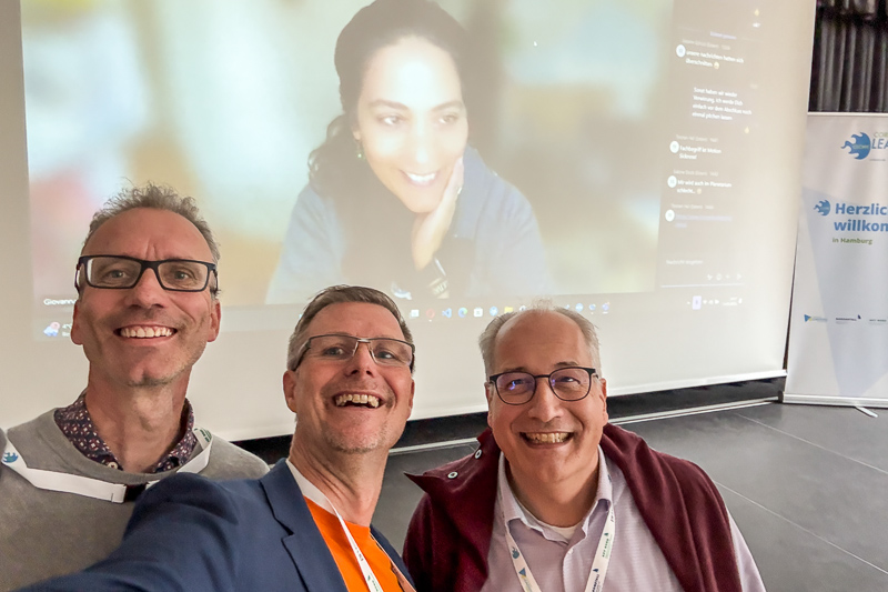 Die CLCFour an Tag 2 ganz hybrid (Quelle: Harald Schirmer), Auf dem Bild stehen drei Männer lächelnd vor einer Leinwand. Es sind
Martin Geisenhainer, Harald Schirmer und Herwig Kummer. Auf der Leinwand
ist eine Frau, Giovanna Lo Presti, in einer Videokonferenz zu sehen --
sie lacht freundlich.