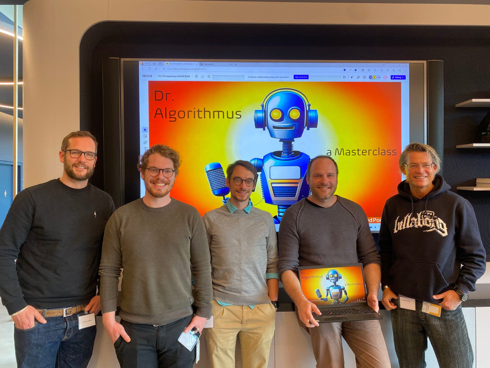 Podcasting-Team (von links nach rechts: Stephan Barber, Torben Linnhoff, Felix Fechner, Rolf, Müller, Torsten Schmidt) auf dem CLC Promptathon (Quelle: Felix Fechner), Auf dem Bild sehen wir fünf Männer nebeneinander, die in die Kamera lächeln. Es sind Stephan Barber, Torben Linnhoff, Felix Fechner, Rolf, Müller und Torsten Schmidt. Hinter ihnen ist ein großer Bildschirm. Auf dem Bildschirm ist ein bunter Hintergrund in Orange und Gelb zu sehen. Darauf sieht man einen blauen Roboter mit Kopfhörern und einem Mikrofon. Außerdem steht dort der Text: „Dr. Algorithmus - a Masterclass".