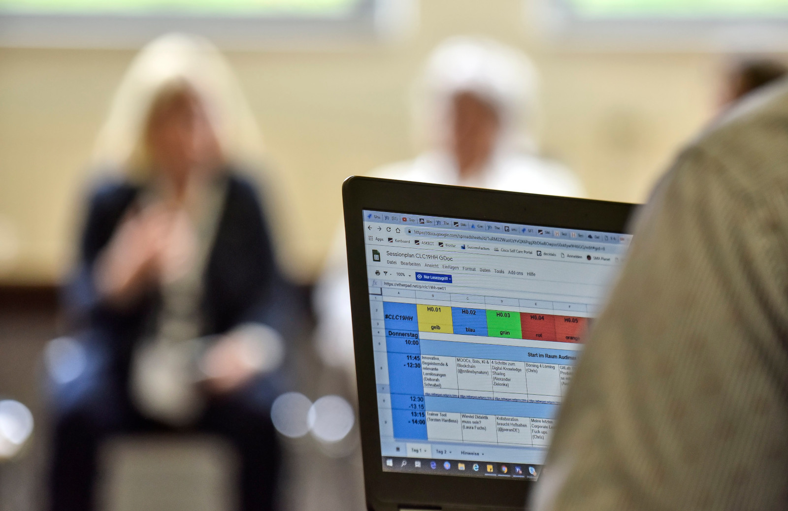 Digitaler Sessionplan auf dem #CLC19HH (Foto von Christian Augustin, im Auftrag von Nordmetall, CC BY 4.0), Das Bild stammt vom Corporate Learning Camp 2019. Auf dem Bild sieht man im Vordergrund einen Laptop. Auf dem Bildschirm ist eine bunte Tabelle mit Terminen und Text zu sehen, ein Ausschnitt aus dem Sessionplan. Im Hintergrund sitzen mehrere Personen und sprechen miteinander, aber sie sind unscharf.