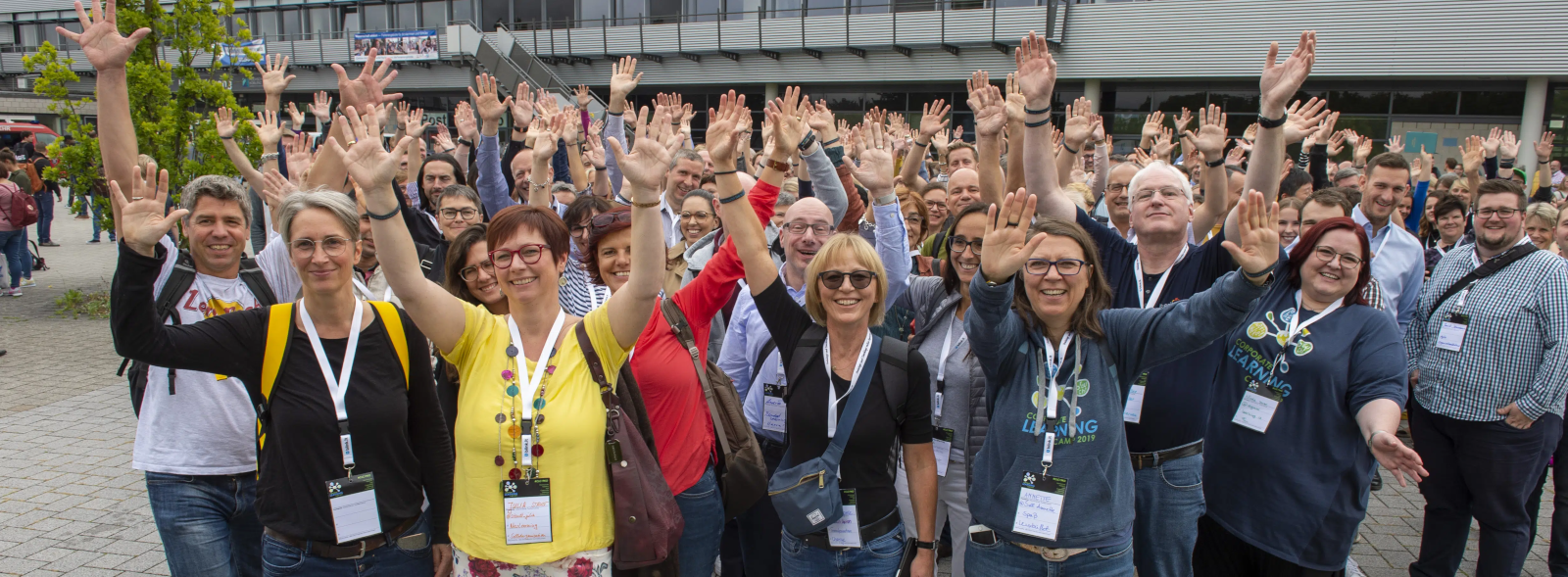Teilgebende auf dem #CLC19 (Foto von Charlotte Venema), Das Foto ist auf dem Corporate Learning Camp 2019 entstanden. Wir sehen eine große Gruppe von Menschen draußen vor einem Gebäude. Viele von ihnen lächeln, schauen in Richtung Kamera und heben beide Hände in die Luft. Viele tragen Namensschilder um den Hals. 
