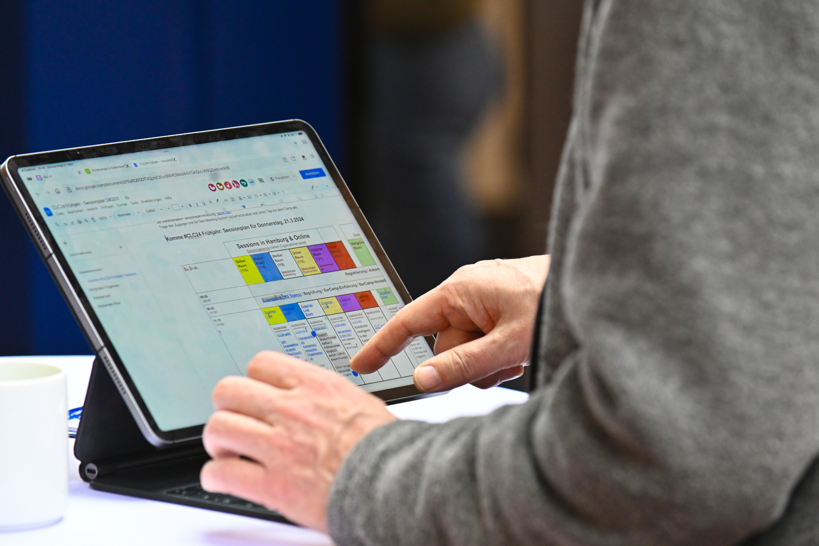 Sessionplan auf dem CLC Barcamp (Quelle: Nordmetall 2024, CC-0), Auf dem Bild sieht man eine Person, die ein Tablet benutzt. Auf dem Bildschirm ist ein bunter Sessionplan eines BarCamps mit verschiedenen Themen und Uhrzeiten zu sehen. Die Person tippt mit dem Finger auf den Bildschirm. Links daneben steht eine weiße Tasse.