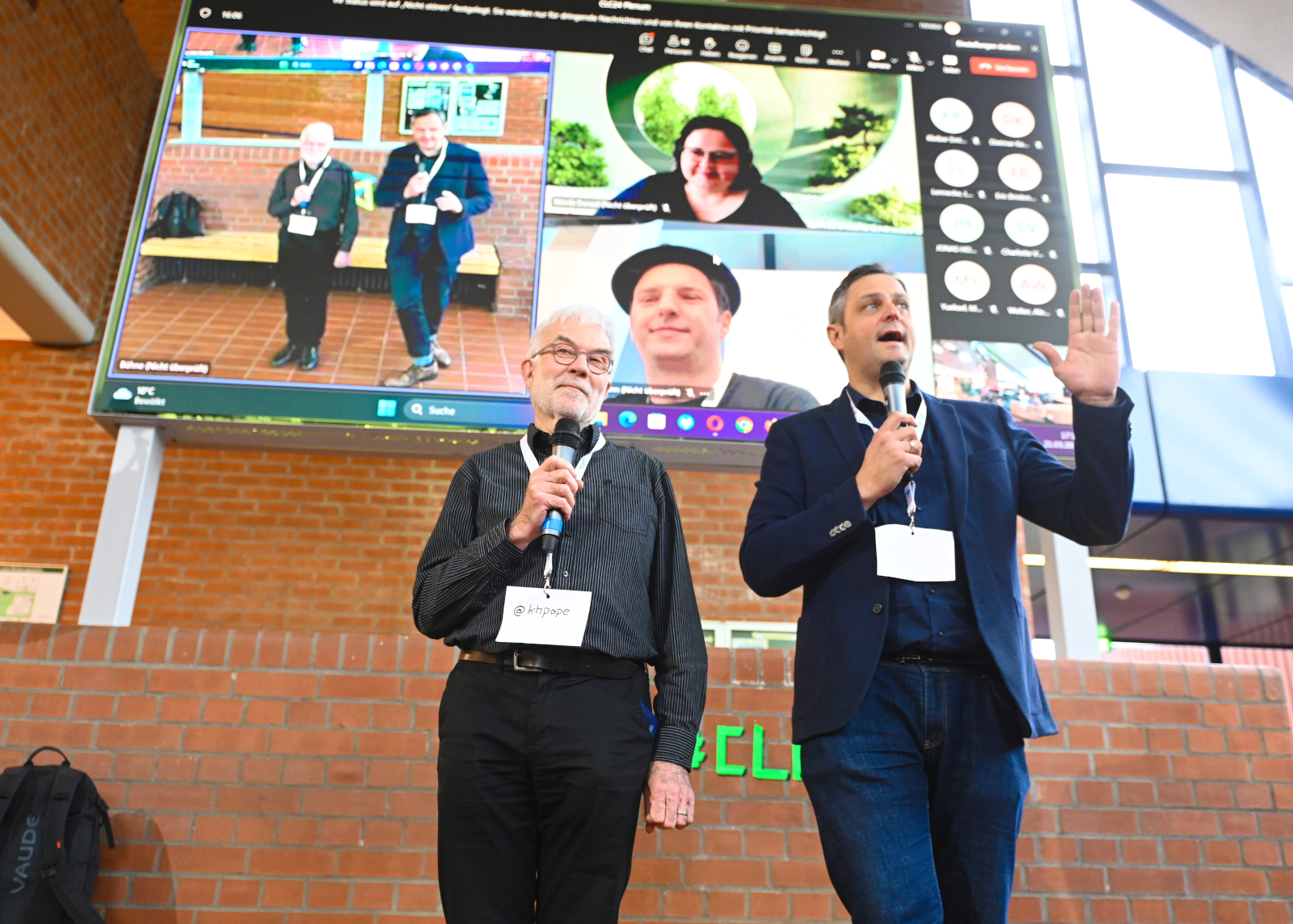 Karlheinz Pape und Henning Klaffke. Moderation auf dem hybriden Barcamp in Hamburg, 2024 (Quelle: Nordmetall 2024, CC-0), Auf dem Bild stehen zwei Männer mit Mikrofonen auf einer Bühne. Es sind Karlheinz Pape und Henning Klaffke, die das Corporate Learning Camp 2024 in Hamburg moderieren. Sie tragen Namensschilder und sprechen zu einem Publikum. Hinter ihnen ist ein großer Bildschirm zu sehen. Darauf läuft eine Videokonferenz mit mehreren Teilnehmenden, deren Gesichter in kleinen Fenstern angezeigt werden.