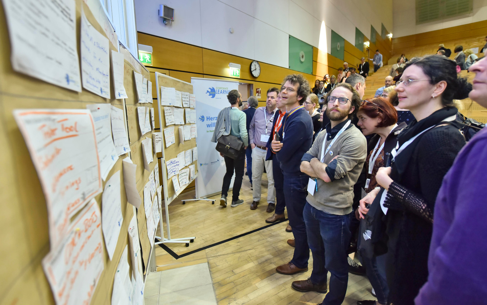 Andrang vor dem Sessionplan auf dem #CLC19HH (Foto von Christian Augustin, im Auftrag von Nordmetall, CC BY 4.0), Das Bild ist auf dem Corporate Learning Camp 2019 in Hamburg entstanden. Auf dem Bild stehen viele Menschen vor dem Sessionplan des BarCamps in einem Veranstaltungsraum. An der Wand hängen viele Zettel mit handgeschriebenen Themen. Die Teilnehmenden schauen sich die Vorschläge an. 