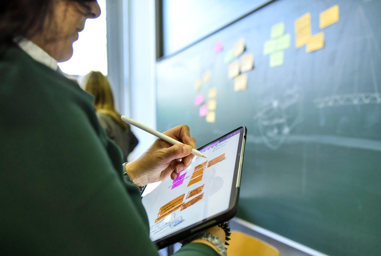 Sketchnoting auf dem #CLC19HH (Foto von Christian Augustin, im Auftrag von Nordmetall, CC BY 4.0), Das Bild stammt vom Corporate Learning Camp 2019 in Hamburg. Auf dem Bild sieht man eine Person, die mit einem digitalen Stift auf einem Tablet schreibt oder zeichnet. Im Hintergrund hängen bunte Zettel an einer großen grünen Tafel. Eine weitere Person steht davor.