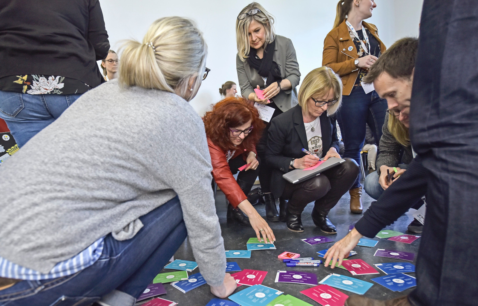 Arbeit in einer Session auf dem #CLC19HH (Foto von Christian Augustin, im Auftrag von Nordmetall, CC BY 4.0), Das Foto stammt vom Corporate Learning Camp 2019 in Hamburg. Auf dem Bild sind mehrere Menschen zu sehen, die sich in einem Raum aktiv an einer Gruppenarbeit beteiligen. Einige knien oder hocken am Boden und sortieren bunte Karten mit Symbolen und Texten. Andere schreiben Notizen oder beobachten das Geschehen.