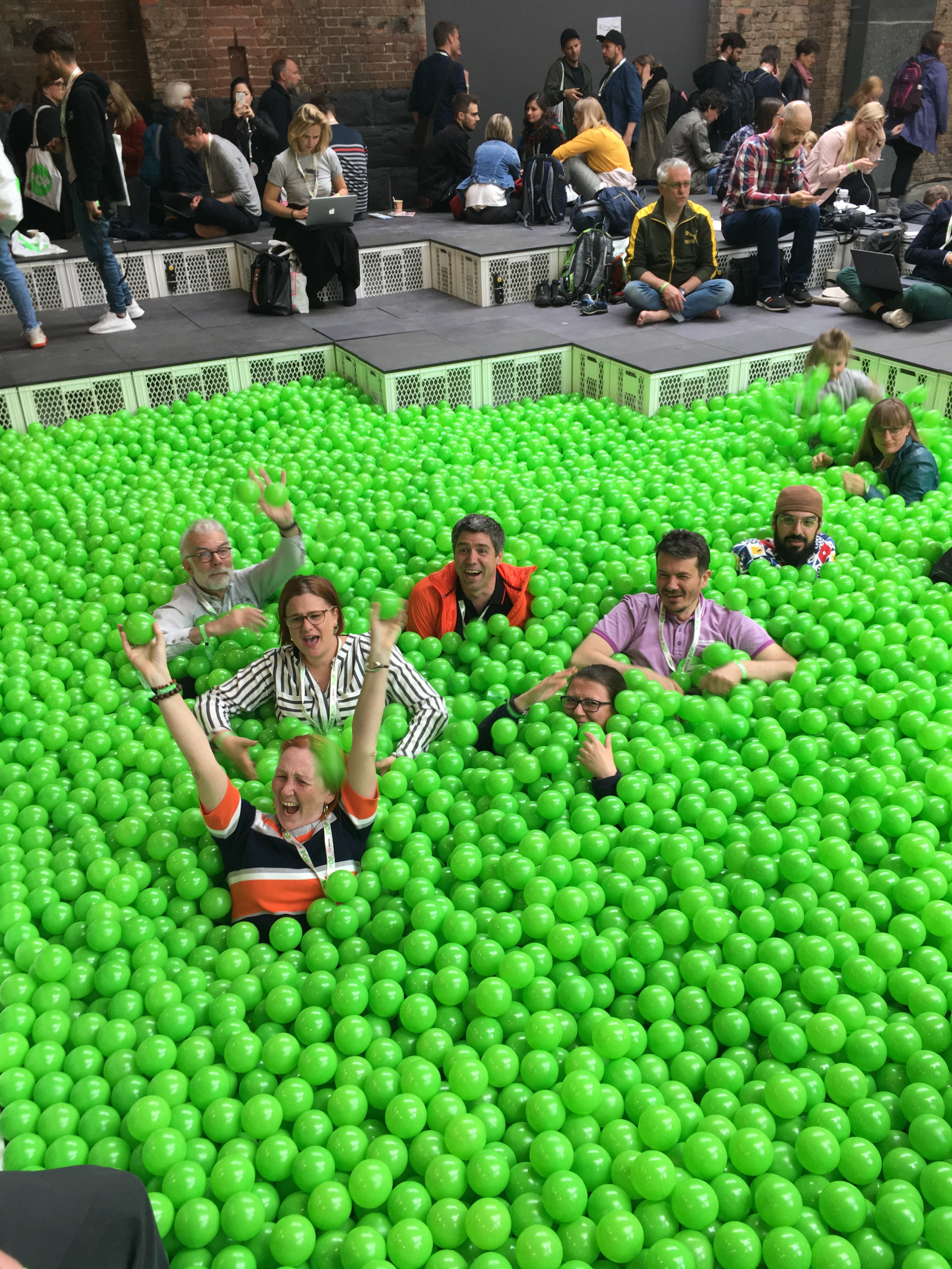 Auf dem Bild sieht man eine große Gruppe von Menschen bei einer
Veranstaltung. Im Vordergrund sitzen mehrere Erwachsene fröhlich in
einem Becken voller grüner Plastikbälle -- wie in einem Bällebad. Sie
lachen, winken und haben sichtbar Spaß. Im Hintergrund sitzen weitere
Personen auf einer Tribüne. Einige arbeiten an Laptops oder unterhalten
sich. 