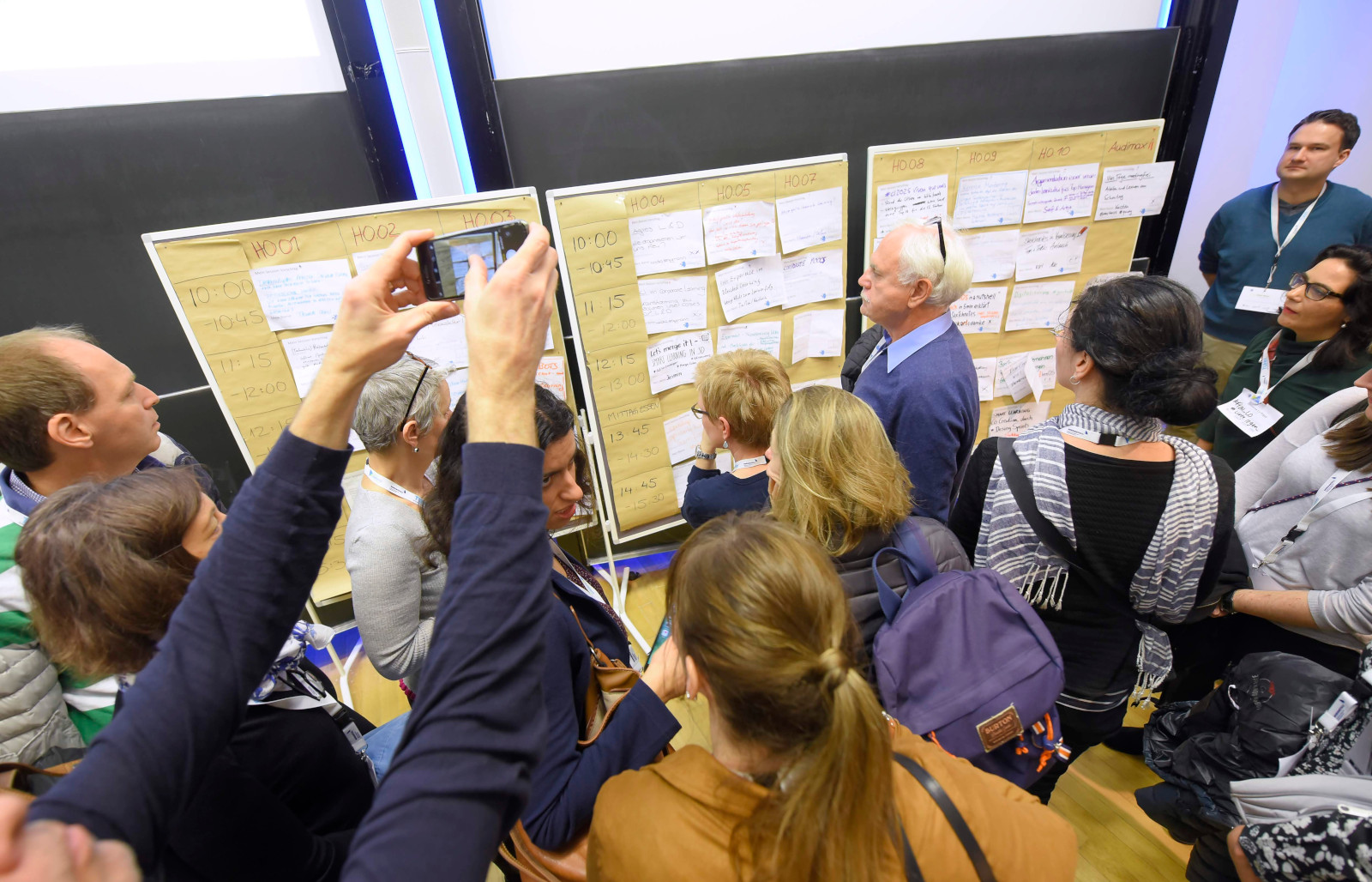 Andrang vor dem Sessionplan auf dem #CLC19HH (Foto von Christian Augustin, im Auftrag von Nordmetall, CC BY 4.0), Das Bild stammt vom Corporate Learning Camp 2019 in Hamburg. Auf dem Bild stehen viele Menschen dicht gedrängt vor einer großen Pinnwand. An der Wand hängen viele Zettel mit Uhrzeiten, Themen und Raumnummern. Es ist der Sessionplan des BarCamps. Einige Personen lesen die Zettel, andere fotografieren den Plan mit dem Handy.